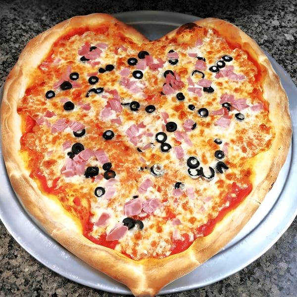 Heart-shaped Zio's Pizzeria pizza with ham and black olives on a metal tray.