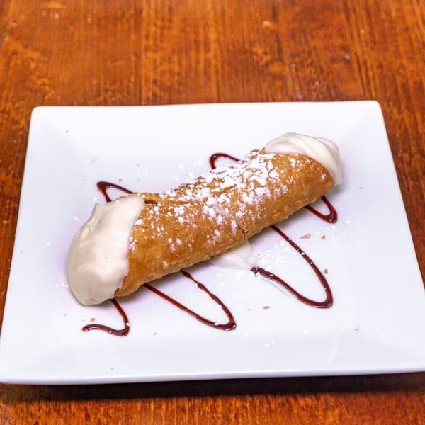 A Zio's Pizzeria cannoli, dusted with powdered sugar and drizzled with chocolate, on a white plate.