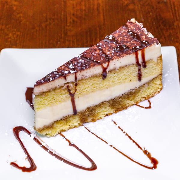A slice of tiramisu cake with chocolate drizzle and powdered sugar on a white plate.
